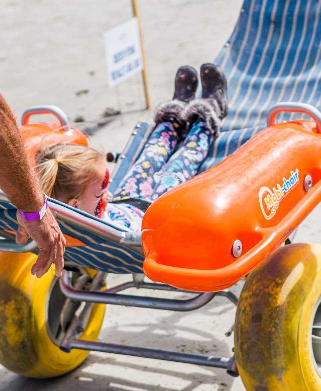 A young girl is pushed on the beach in a large-wheeled wheelchair