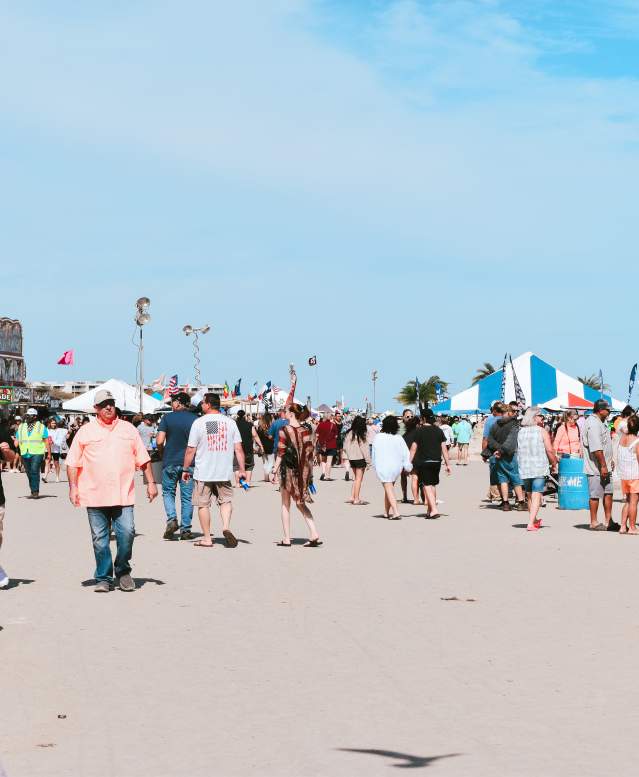 A crowd of people walking along the beach with carnival tents