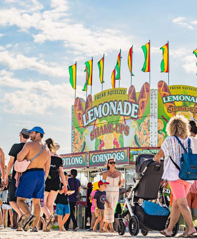 A crowd of people on the sand pass by a large stand selling lemonade and food items