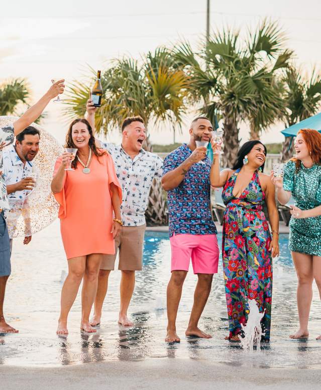 A group of eight people, a mix of men and womenm, stand ankle deep in a pool and celebrate with champagne