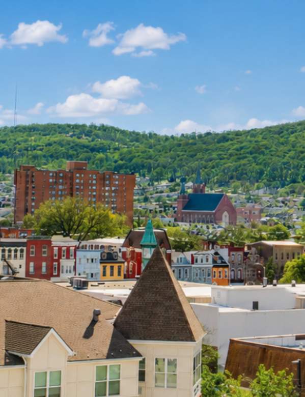 Panoramic summer view of a small city surrounded by green, tree-covered hills under a bright blue sky with scattered white clouds. In the foreground are colorful apartment buildings and rooftops, while brick high-rise residential towers rise above the downtown area. Tree-lined streets and historic church steeples are visible throughout the cityscape, creating a vibrant, sunny urban scene.