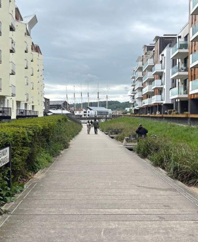 An image of a wide pathway with neat green bushes running along either side. There are tall buildings on either side that are light yellow and clad in wood. In the distance there are two people walking ahead and even further there are six masts of a ship.