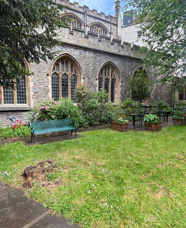 An image of a grassy lawn area surrounded by narrow paved paths. There is a church behind the path and benches and flowers pots on the path running alongside the church.