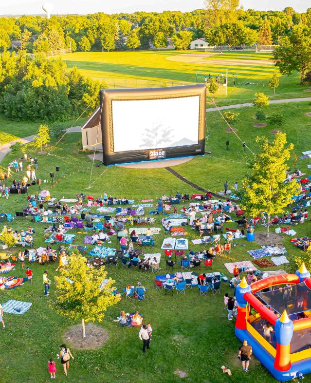 Outdoor movie screen at Ojibway Park with movie-goers seated on a hill with bounce house in the foreground.
