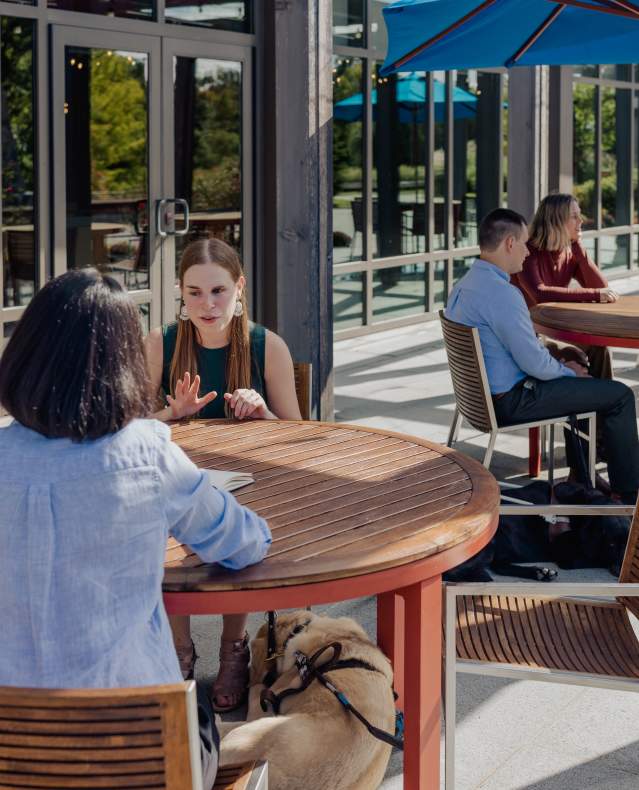 Three women sit together outside chatting at a table. A guide dog is sleeping at the feet of one of the women.