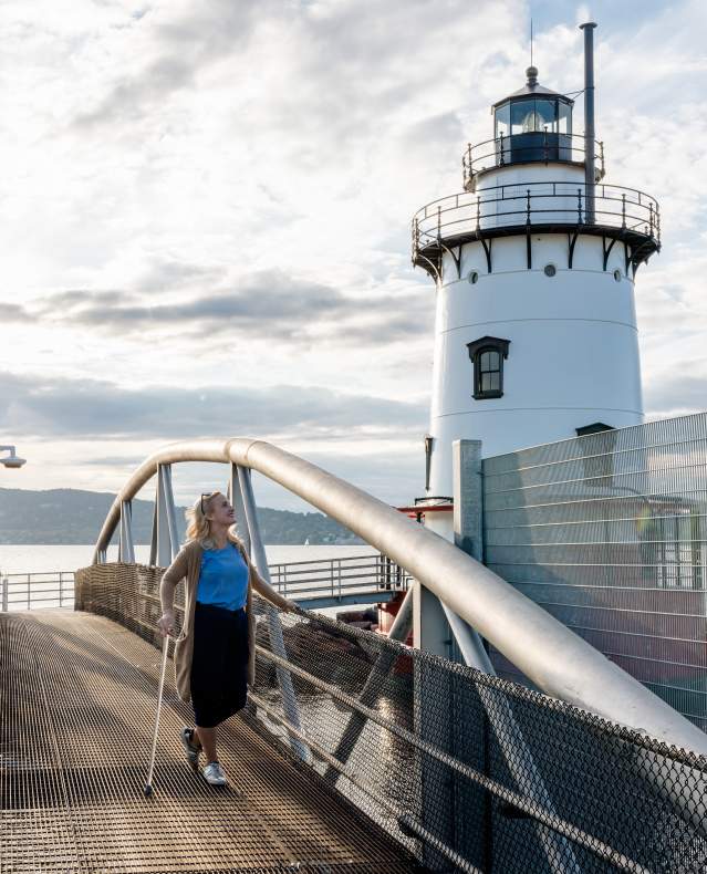 A woman with a cane stands on the bridge to the Tarrytown Lighthouse. She is looking out on the Hudson River and smiling.