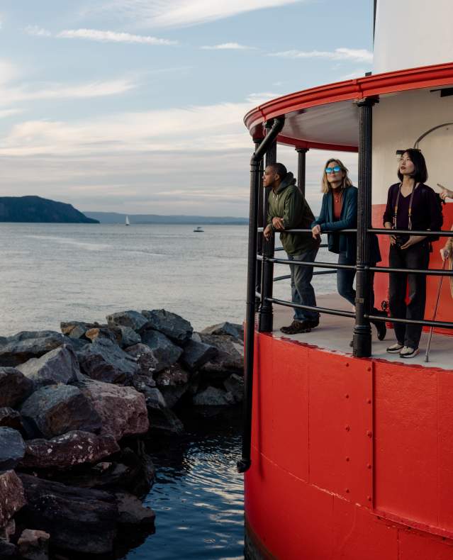 Five people stand on the balcony of the Tarrytown Lighthouse at sunset looking out at the Hudson River.