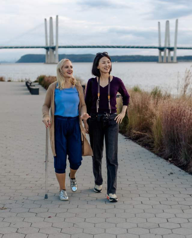 Two people are walking along the Riverwalk in Tarrytown. The Gov. Mario Cuomo Bridge is in the background.