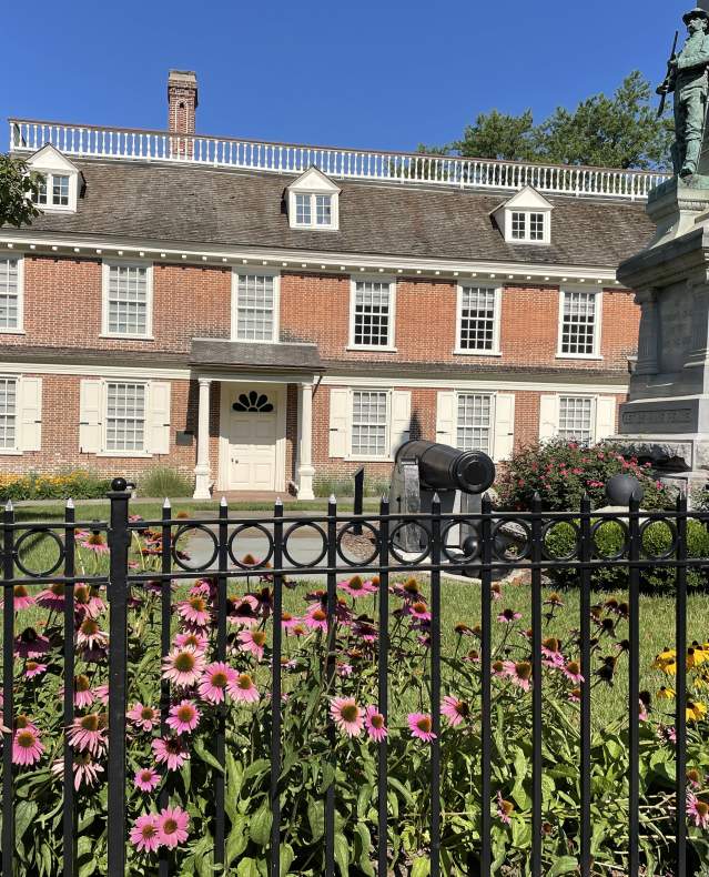 The exterior of Philipse Manor Hall with purple and yellow flowers blooming in the foreground.