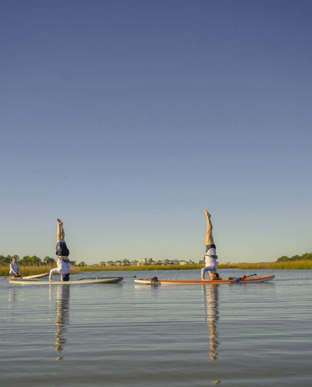 Women practicing paddleboard yoga with headstands on calm marsh waters in North Myrtle Beach, South Carolina, under a clear blue sky surrounded by peaceful natural scenery.