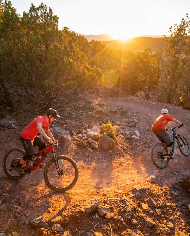 Two cyclists rounding a turn on the Lichen It mountain bike trail in Cedar City Utah at sunset with a sunburst over the distant hills.