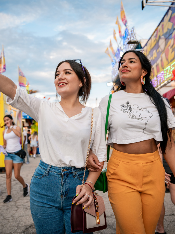 Two young women walk arm-in-arm through the Wisconsin State Fair, with one pointing excitedly ahead. Carnival rides, game booths, and crowds create a festive atmosphere in the background.