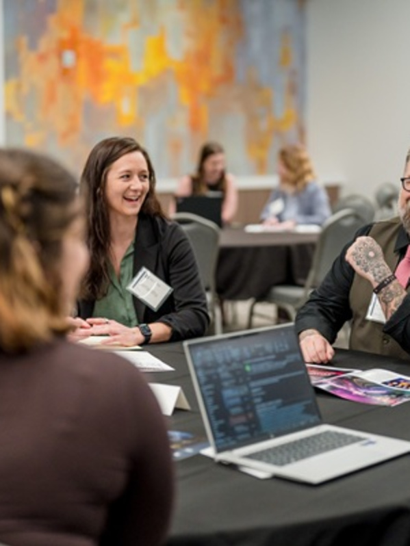 Attendees participate in a networking session around a round table at a conference. Several people smile and chat while reviewing materials and a laptop, creating a collaborative, professional atmosphere in a meeting room.