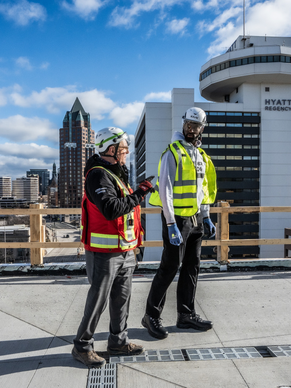 two people in construction gear on an outdoor terrace with the Milwaukee skyline behind them