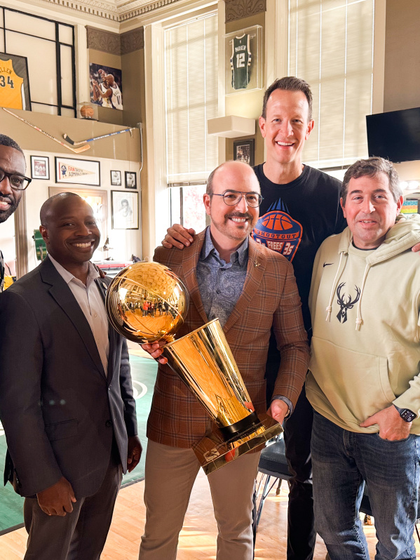 A group of five men pose together at Gee's Clippers. From left to right: Gee in a black and white striped referee shirt, Mayor Johnson in a dark business suit, David Caruso in a brown plaid blazer holding a golden basketball championship trophy,  Steve Novak in a dark shirt, and Peter Feigin in a cream-colored hoodie with a deer logo. The room features basketball memorabilia on the walls including framed jerseys and photos, a basketball hoop, and large windows.