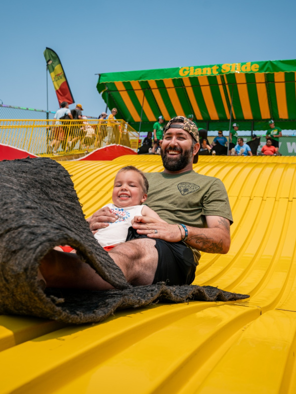 A smiling man holds a young child as they ride down the bright yellow Giant Slide together on a burlap mat at the Wisconsin State Fair.