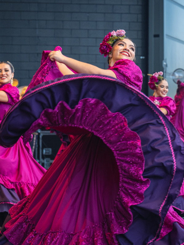 A group of female dancers performs a traditional Mexican folklórico dance on stage. They wear vibrant magenta dresses with purple ruffles and floral headpieces. The lead dancer in the foreground smiles brightly while gracefully twirling her flowing skirt. The dancers behind her mirror the movement, creating a colorful, energetic scene against a modern stage backdrop.