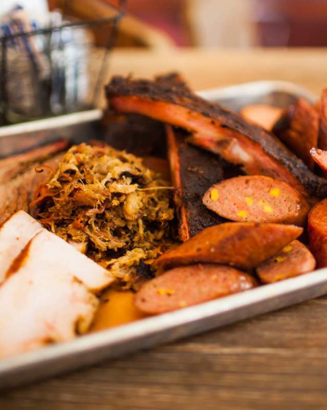 Plate of barbecue food from Backdoor BBQ featuring ribs, pulled pork, smoked turkey and more. The tray is sitting on a wooden table.