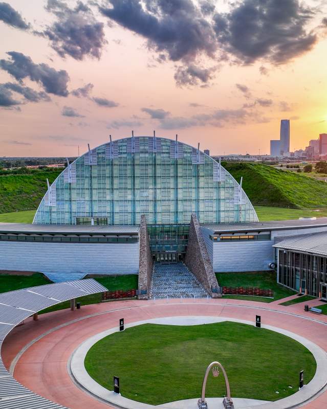 Drone shot of the First Americans Museum at dusk, in the background is the downtown OKC skyline