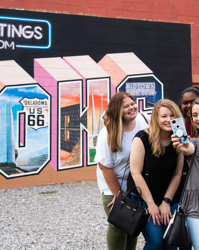 Four women posing for a selfie in front of Midtown's "Greetings from OKC" wall mural