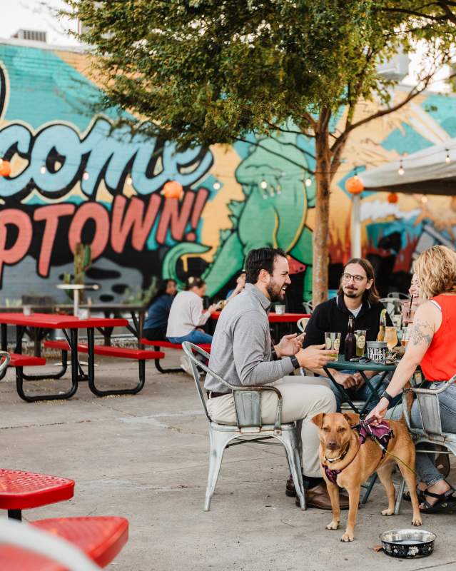 Three people have drinks with their dog on the patio at Pump Bar. You can see a mural in the background that says, "Welcome to Uptown".
