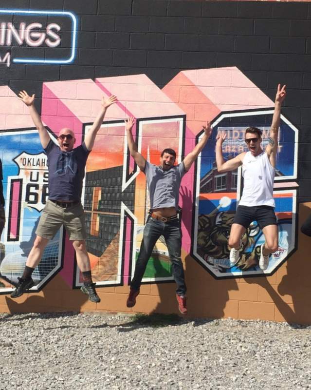 Photo of five men jumping for joy in front of the "Greetings From OKC" mural.