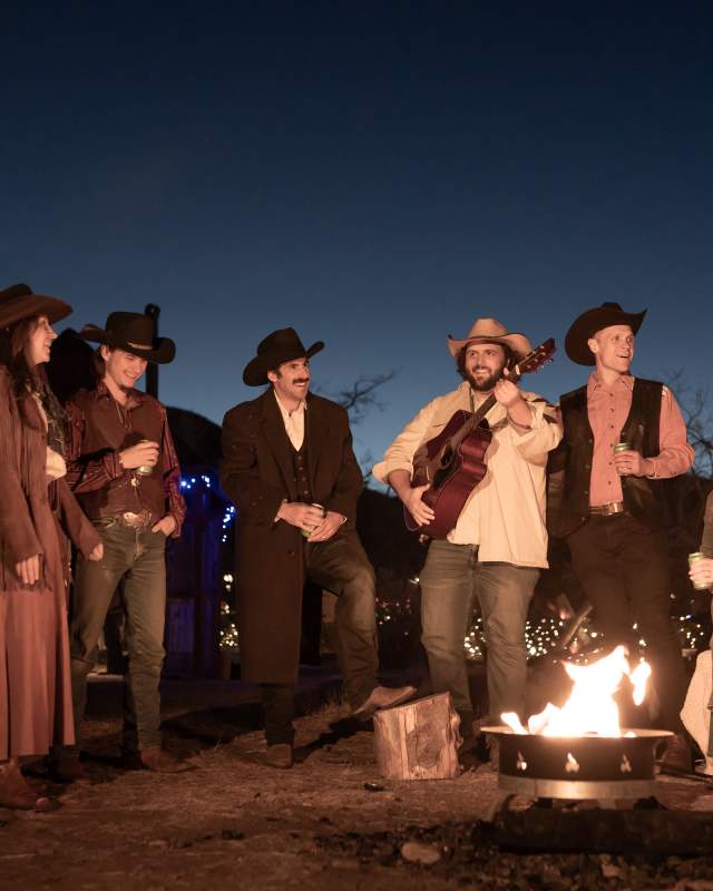 People around a campfire in Golden in western attire, one person is playing a guitar
