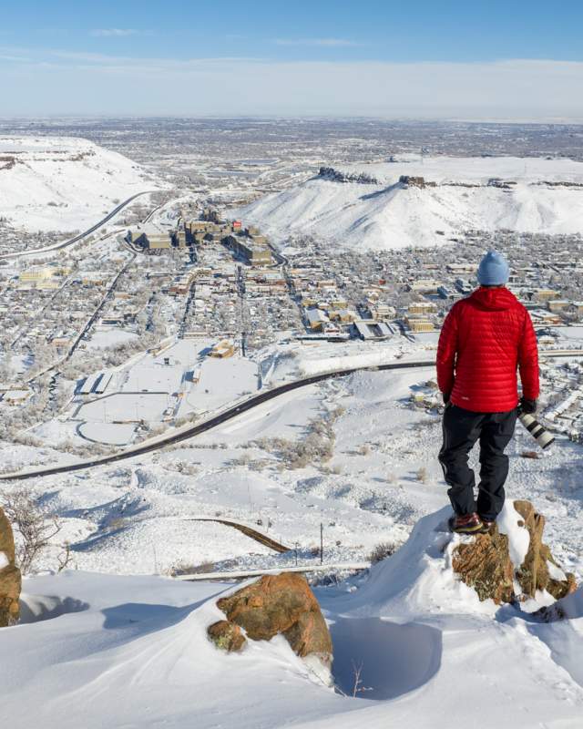 Person in a red coat looks out over snow covered Golden from high above on a sunny day from Lookout Mountain