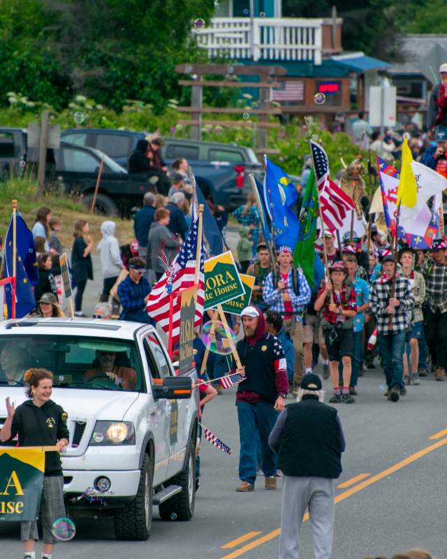 4th of July Parade in Homer, Alaska with white car leading a group of people marching down the street with various flags.