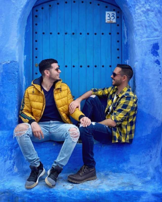 2 men holding hands and sitting in front of a blue door and wall