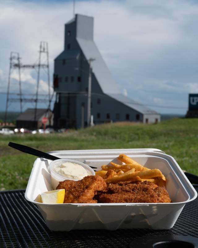 a to go container full of fried fish and french fries sits on an outdoor table in sight of the quincy mine