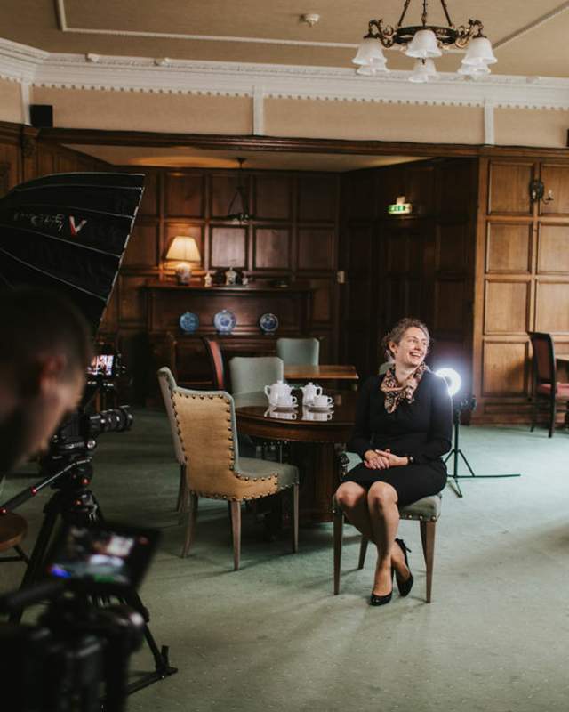 A woman sat on a chair in the middle of a hotel lounge with camera and a cameraperson in the foreground