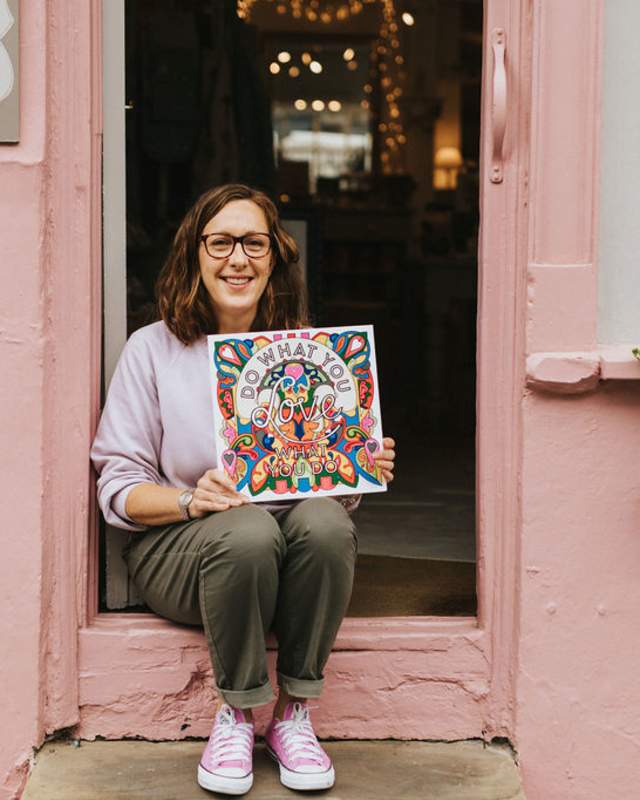 Woman sat outside her gift shop holding a shop local sign