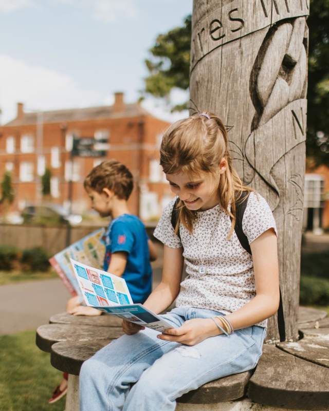 Girl looking at a treasure trail map sat on a bench with young boy in background