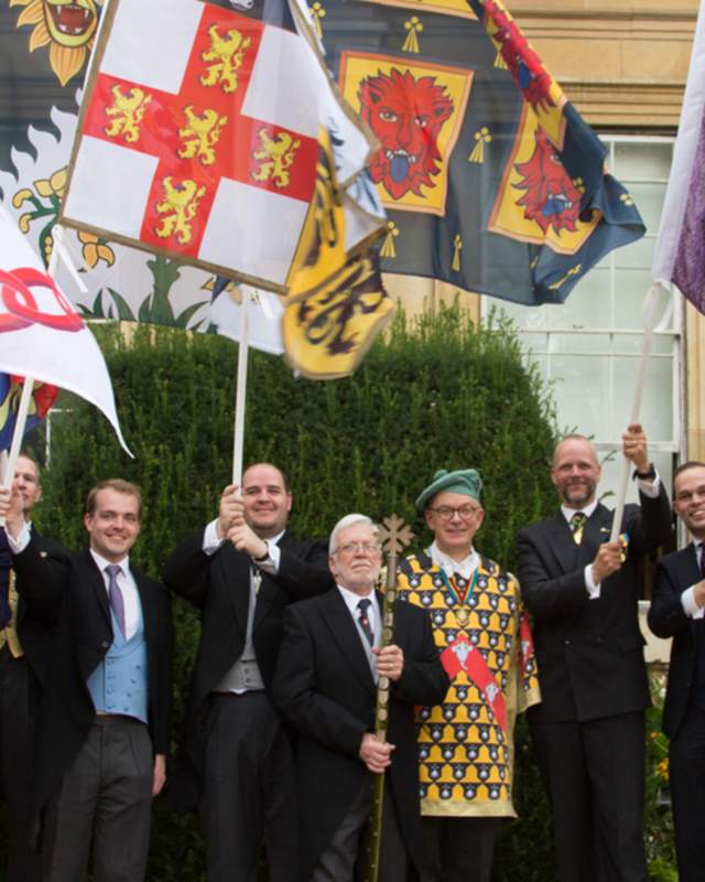 Delegates at the 35th International Congress of Genealogical & Heraldic Sciences stand in a group wearing uniforms and holding heraldic flags.