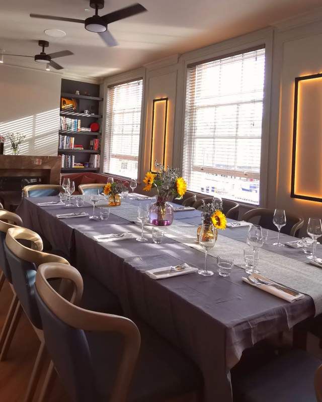 Dining table set up for private dining at Market House Cambridge. The table has a dark tablecloth with white place settings & posies of flowers in the centre of the table/ The room is light & bright with large windows to the right of the image