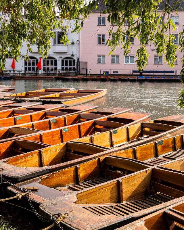 A row of 12 punts on the river in Cambridge with a stone bridge to the left & a white & pink building on the other side of the river