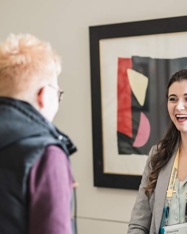 A young white woman with long wavy brown hair smiling in front of an abstract art picture whilst chatting to another white woman who has her back to the camera