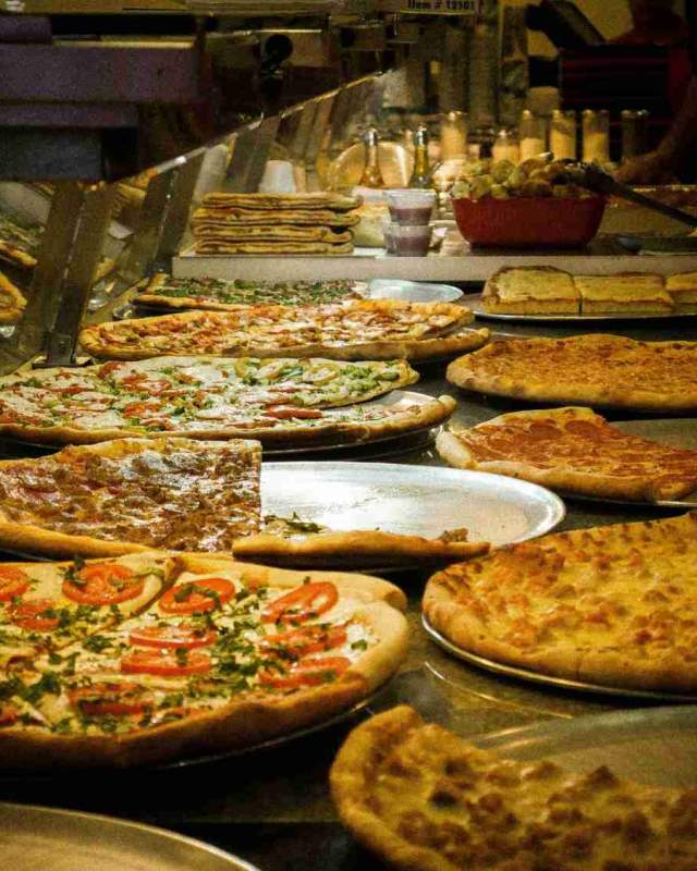 Fresh New York pizza slices displayed behind the counter at a busy Manhattan pizzeria with classic thin-crust and Sicilian-style pies.