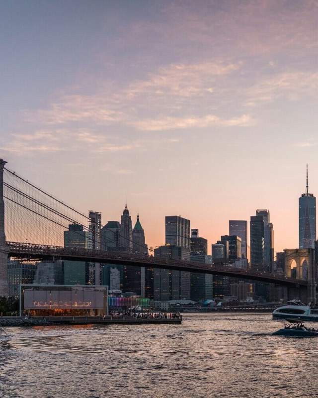 Brooklyn Bridge with the Manhattan skyline glowing behind it at sunset, viewed from the water in New York City