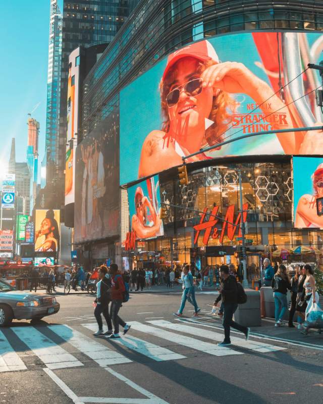Busy daytime intersection in Times Square, New York City, with pedestrians crossing, large digital billboards, and the H&M storefront surrounded by tall buildings.