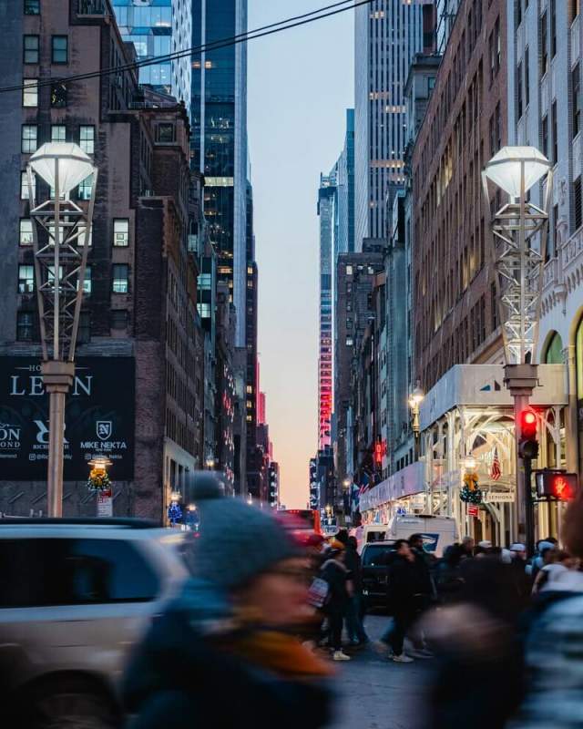 Blurred pedestrians crossing a busy Midtown Manhattan intersection at dusk with skyscrapers and red traffic lights