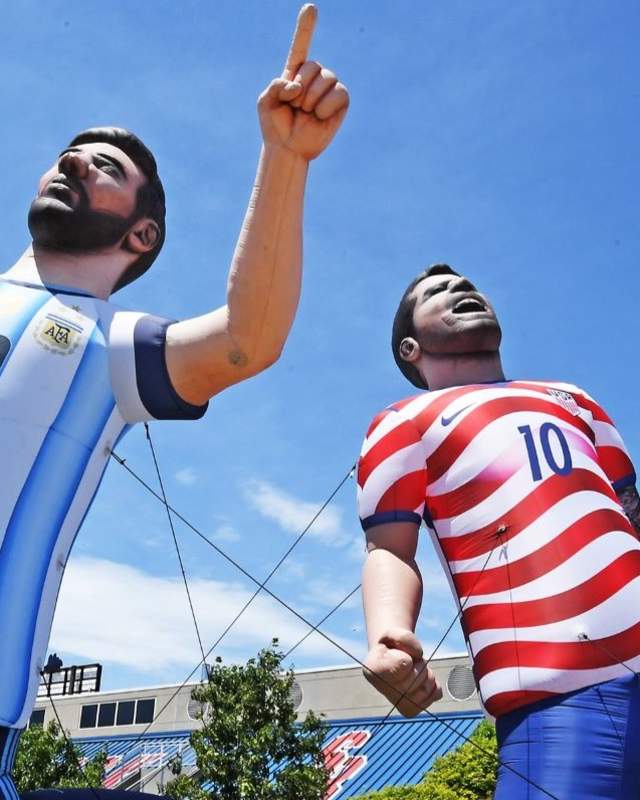 Two large inflatable soccer players kneeling, one in an Argentina jersey, the other in a USA jersey, with a giant ball nearby. Clear blue sky backdrop.