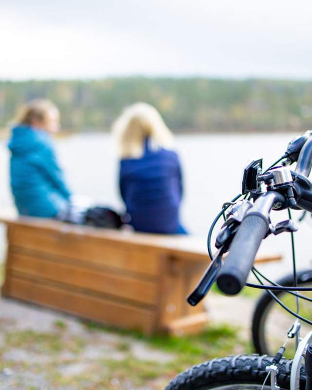 To jenter sitter på en krakk og ser ut mot havet. Syklene deres står i forgrunnen, og er i fokus.