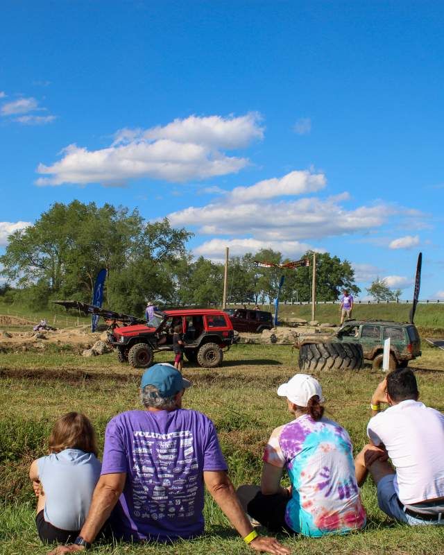 A group of six people sitting on grass in a field watching two jeeps race through an obstacle course