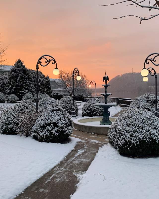 A quaint fountain in a park with snow covered ground and greenery surrounding and a sunset in the background at Danville's Riverfront