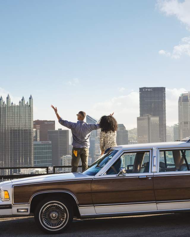 Couple stands behind car overlooking a city skyline of Pittsburgh, Pennsylvania