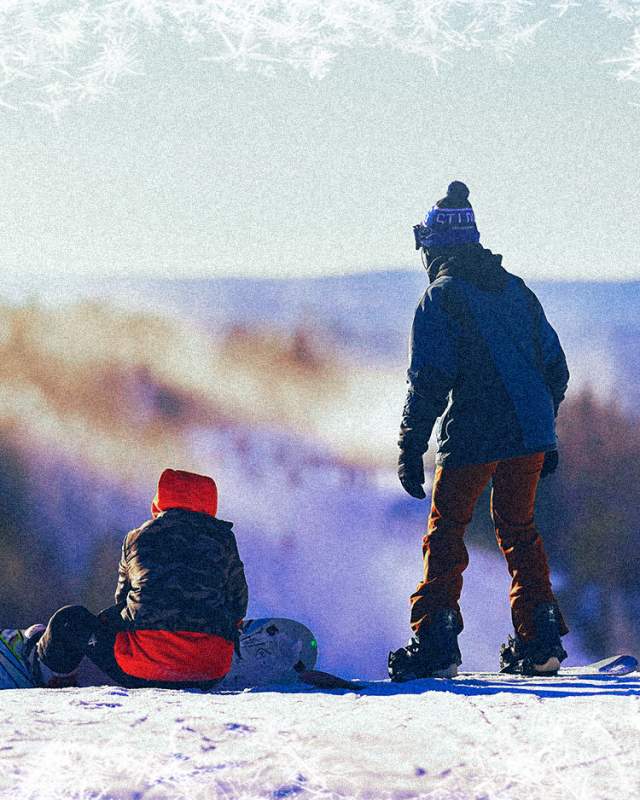Two kids in snow gear on a snowy mountain. One is sitting and the other is standing on a snowboard.