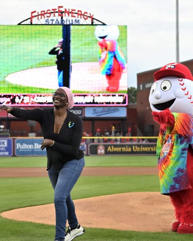 A person throws out the first pitch at the baseball game with Screwball (the R Phils mascot) cheering her on in a tie dye shirt.