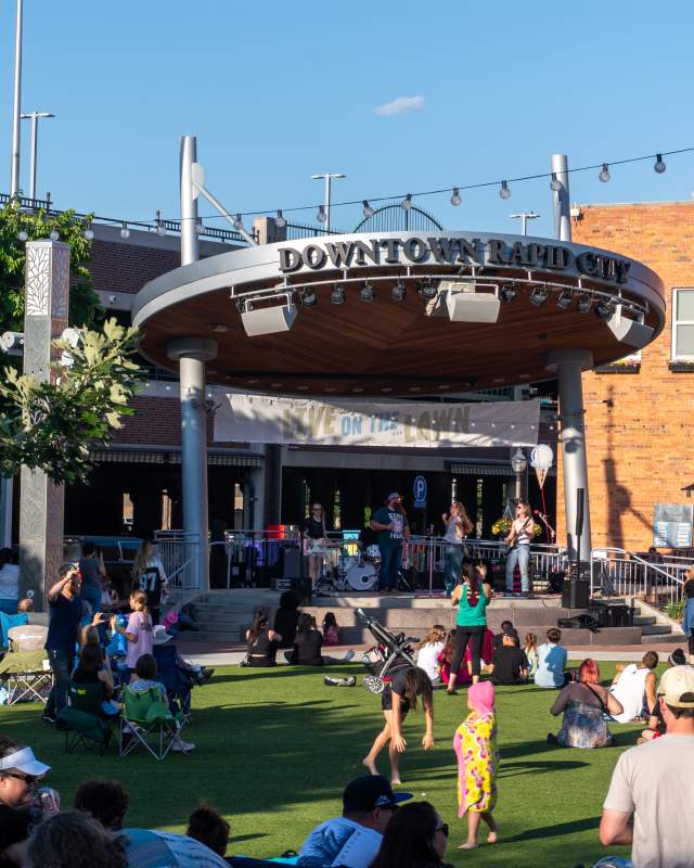 Looking at a distance towards a band playing on the stage at Main Street Square while attendees sit in lawn chairs scattered throughout the grass.